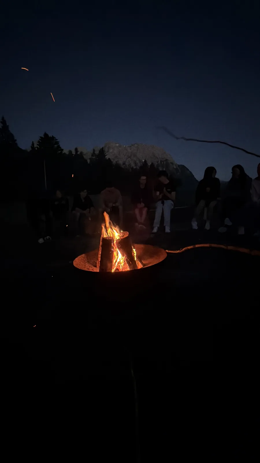 TUM.ai-Mitglieder versammeln sich in der Dämmerung um ein Lagerfeuer, dahinter schemenhaft die Berge.