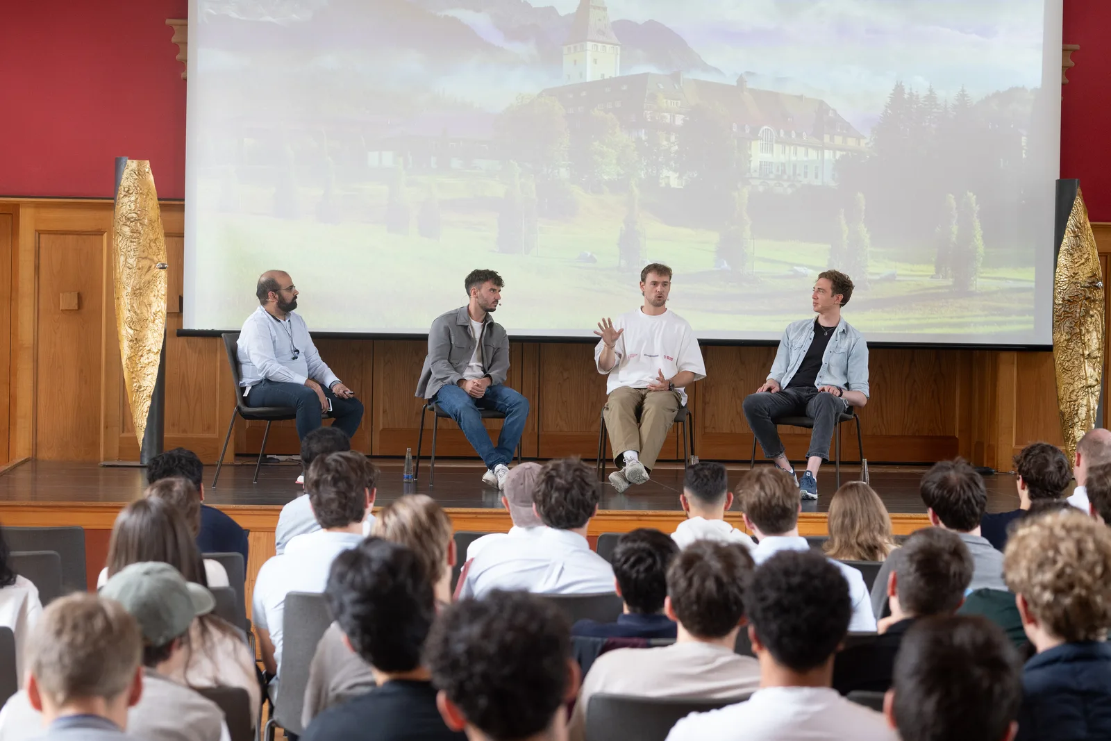 Vier Personen sitzen auf einer Bühne vor einem großen projizierten Bild von Schloss Elmau und sprechen zu einem sitzenden Publikum.